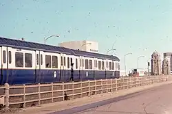 A metro train on an urban bridge. The cars are dark blue with a white stripe over the windows.