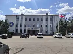 A large, gray administrative building with a parking lot in the foreground. To the right of the building, the flags of Georgia and the European Union are flying on flagpoles. Shot on a sunny day against a blue sky with white clouds.