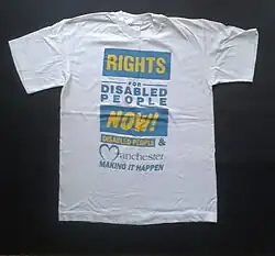 This is a white t-shirt on a black background. The lettering is blue on white and yellow on blue. It says, "Rights for Disabled People Now!" and "Disabled People & Manchester Making It Happen".