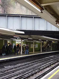 Eight people walking towards an escalator on a railway platform next to a green-tiled wall on the left and a railway track on the right