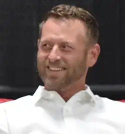 A 42-year-old man with brown hair and a brown beard smiling to the left of the camera.