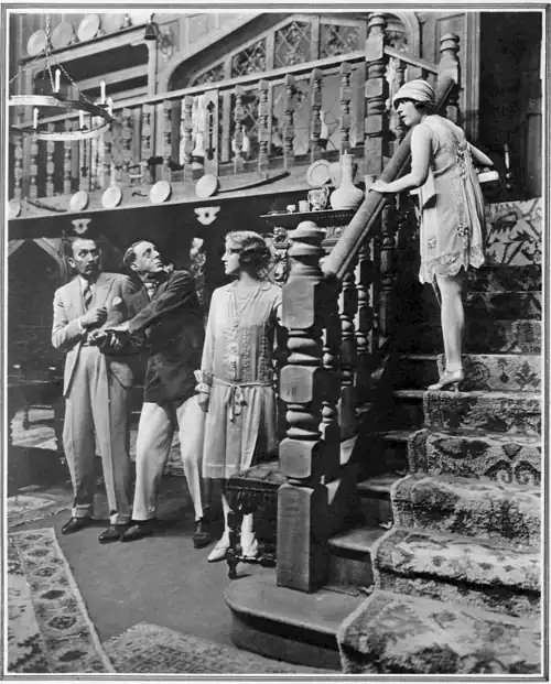 interior of country house with two men and a woman, standing, ground level and a young woman half-way up the stairs to the gallery