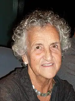 Portrait photograph of smiling, gray-haired woman wearing a brown V-neck shirt and a silver necklace.