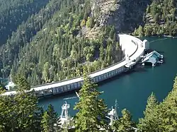 A curved concrete dam, viewed from slightly above. On the left is a steep forested valley, and on the right is blue-green water. The reservoir is almost full.