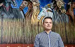 The image shows Aaron Paquette standing in front of a painting that takes up the entire background of the frame. It depicts horses in a war paint in a textured and gestural painting style but with additional finer details. Vibrant colours and movement. The horses are standing in front of a night sky in shades of blue and behind a “wall” of sweetgrass that extends along the lower quarter of the painting. Beneath that in the lowest part of the painting is a red stripe.