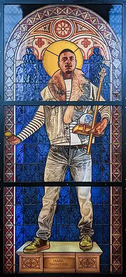 Stained glass window in a romanesque arch shape. A young black man with a golden halo, wearing modern clothes, standing on a white and gold plinth inscribed "Mark Shavers". He is holding an egg in one hand, and a book, orb and scepter in the other hand. The background is a deep blue, and the window is framed with geometric patterns