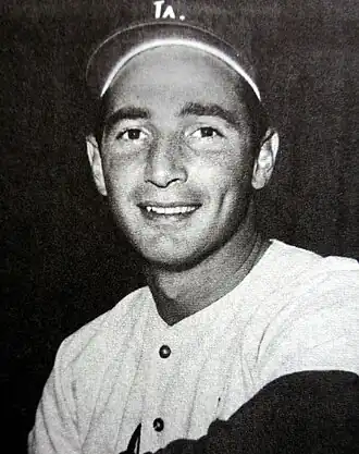 Black and white photo of a man in a white baseball jersey and dark baseball cap with an interlocked white "LA" on the front, smiling at the camera.