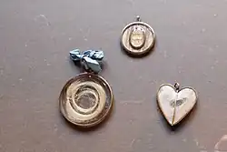 In this photo there are three fancy lockets, they are small and ornate, likely they are from a museum or private collection from the fancy photo and cloth they're on. None of them have chains attached so they are probably old and not currently being worn. They are made of precious metals. Two contain a light color of hair, either sunbleached, blonde, or greyed with age.