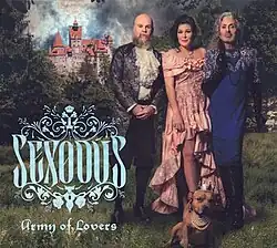 All 3 Army of Lovers members standing in a grassy forest clearing, with a castle and ominous sky in the background. Alexander is wearing a frilly conductor outfit. Dominika is wearing a pink dress that hikes up around the legs. Jean-Pierre is wearing a blue skin-tight dress with silver accents. There is a small brown dog in front of them, wearing a silver crown and pearl necklace. In the bottom left corner is the album logo and band logo in light blue.