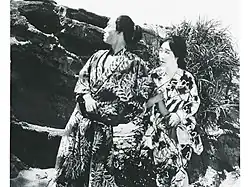 A still from Shima no onna (1920) with Henry Kotani and Yoshiko Kawada; two Japanese actors, a man and a woman, wearing printed kimonos, seated outdoors
