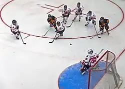 A hockey puck in midair over the ice between a group of five players in white jerseys and two in black at the top of the image, and a goalkeeper in white at the bottom