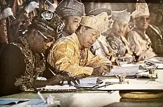 Two men in traditional Malay formal outfits sitting at a table signing their own copies of a document