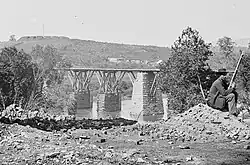 Black and white photo of a bridge, with a seated sentry at right and a fort crowning the height at upper left.