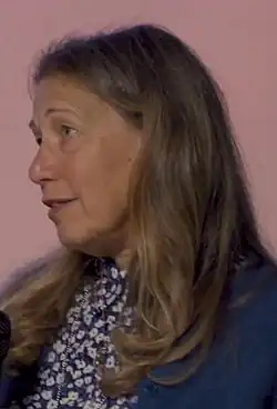 Susan Kare, a woman in a floral print shirt and navy jacket, with long brown hair, looking away from the camera, in front of a pink background