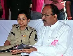 Photograph of a man and a policewoman sitting on a white bench at a celebration. The policewoman, Swati Lakra, is wearing a Hyperabad Police uniform. The man beside her is holding a phone.