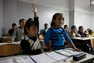 Children at desks in a classroom. One child raises her hand.