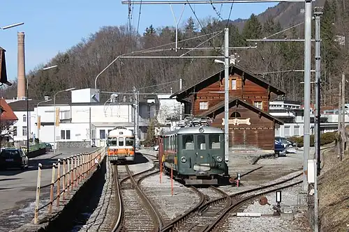 Two electric trains, one green and one orange and white, next to a two-story wooden building