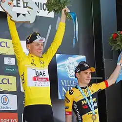 Tadej Pogačar (left) holding Paris-Nice trophy above his head, Jonas Vingegaard (right) holding flowers.