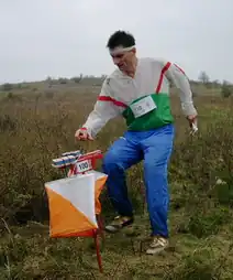 A man in a field with a map in his left hand reaching with his right for a numbered control point marked with orienteering flag.