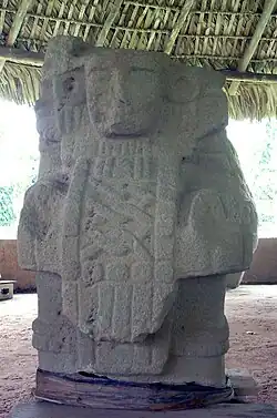 A relief sculpture of a human bust facing forwards, with sculpted cloth hanging over the chest bearing a criss-cross design. The face has a damaged right cheek and two round earspools decorate the ears.