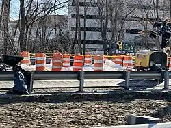 a white tarp covers the second sinkhole, bordered by large traffic cones and caution tape with 2 construction vehicles in the right of the photo and a wheelbarrow on the left, taken from the westbound lanes of the highway