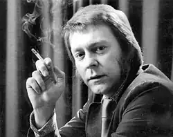 Ted Lewis, is sitting facing the camera. He has a lit cigarette in an outstretched arm and is wearing a suit and tie