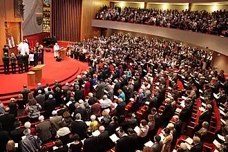 An aerial view of large semi-circular room, filled with rows of curved pews, and a similar balcony on top. People stand in front of most of the seats, holding open books. Many of the men are wearing kippahs (skull-caps). The pews face a raised platform at the front of the room, and in front of that, an open ark containing Torahs. A number of men and women stand in front of the open ark, and above it is a large wall decoration as tall as a person, holding an eternal light.