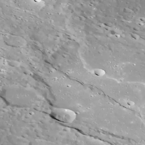 Oblique view of southern Thākur crater. Two prominent lobate scarps deform the crater. These scarps form when one block of crust thrusts up and over another, and are thought to have formed due to a reduction in Mercury's surface area as its interior cooled and contracted. The face of a scarp indicates the direction these blocks have moved so, as we see it here, Tharkur has been squeezed from the top-right and from the bottom-left.[3]