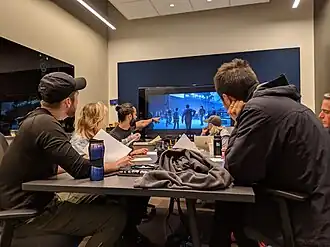 Several people sitting around a table with scripts, looking at a television with some gameplay footage.