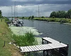 A broad, flat artificial cut with dark water and bright green raised banks. A patch of green algae stains the water. Several modern yachts are moored on wooden jetties spaced along the bank