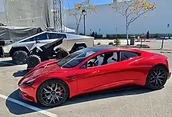 A stainless steel clad ATV between a Tesla Cybertruck and a second generation Tesla Roadster in a parking lot during a Tesla event in 2020.
