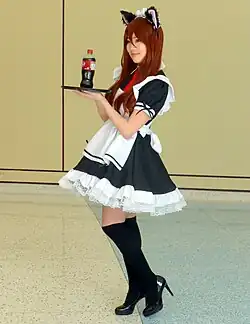 A young woman is wearing a maid outfit and cat ears and presenting a bottle of soda on a tray.