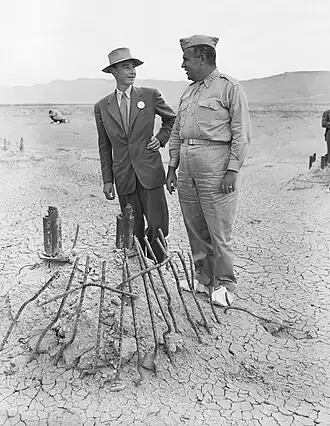 Two men, one in a suit and hat and the other in military uniform, stand in front of twisted metal whilst wearing white overshoes