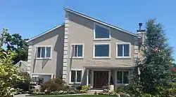 A light brown house surrounded by bushes and trees, with a forklift and an automobile in the driveway, and solar panels on the roof.