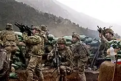 Seven soldiers in beige tactical gear huddle behind a row of green sandbags on a mountainside, pointing rifles in various directions