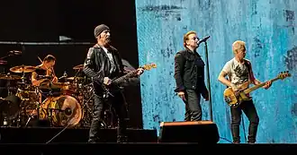 U2 performing on a concert stage. The Edge and Adam Clayton, playing guitars, flank Bono in the foreground, while Larry Mullen Jr. is behind a drum kit in the background on the left side.