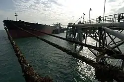 A large red and black tanker ship floats in calm dark waters, tightly moored by ropes to a long steel truss dock, while large pipes pump oil out to the ship