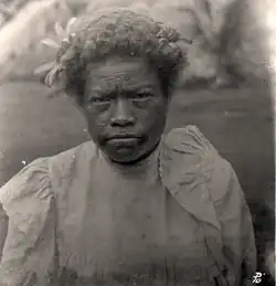 Black-and-white photograph of a Fijian woman facing the camera. She has dark tattoo marks around her mouth. She has a flower in her short Afro and is wearing a Victorian gown in a light colour which comes up to her neck.