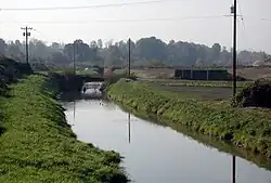 A stream perhaps 20 feet (6.1&nbsp;m) wide flows between grassy banks and telephone poles on either side. Waterfowl float on the stream near a low bridge in the middle distance. In the far distance is a line of trees.