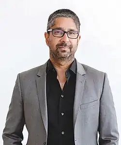 A portrait of Vijay Pande, looking straight ahead. His ethnicity is Indian. He has medium-length black hair, black-rim glasses, and a short mustache and beard. He is wearing a blue polo shirt under a black suit coat.