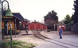 An elderly man standing next to railroad track in the foreground with an old-style railroad depot building on the opposite side. A locomotive shed and water tower are located where the railroad track ends in the background.