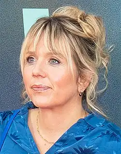 close-up of Kerry Godliman wearing a blue patterned top and gold necklace, looking right of camera with slight smile