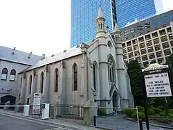 A grey stone church with skyscrapers in the background. The windows have pointed arches, there are two modest pinnacles on the main facade, which has an arched entrance and a traceried window above. A notice board states "Welsh Church", announces forthcoming services, and says "Welcome - Croeso!"