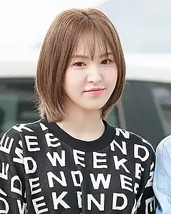 Woman with a short, straight bob haircut and light brown hair. She is smiling gently and wearing a black top with the word "WEEKEND" printed repeatedly in bold white letters.
