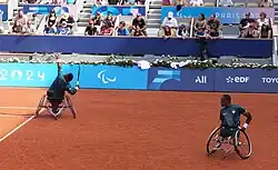 Two Black South Africans play on the same side in a doubles tennis match. Ramphadi, on the left, is in the middle of serving, while spectators watch.