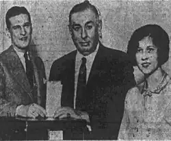 Black and white photograph. Thompson stands before a ballot box, inserting his ballot paper into the box. He has a male. To the left of Thompson is a male, and to the right of him is a woman, both also standing