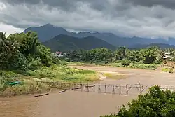 Landscape with a wooden footbridge crossing the Nam Khan river, where 2 workers are working at the consolidation of this structure, holding a beam during the monsoon