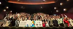 Japanese-language board is presented by smiling people who watched the movie for an event; some in the back have Mario merchandise