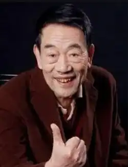 Elderly Chinese man wearing a crimson jacket smiling to the camera in a "thumbs up" pose