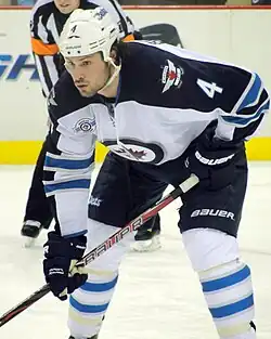 A Caucasian hockey player. He is wearing a white jersey with blue shoulders with a jet over a maple leaf inside of a circle for the logo. He is bent over at the waist leaning on his stick.